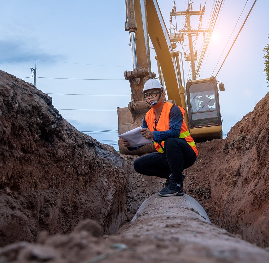 Man in hard hat and safety vest crouches on pipe in trench, reviewing papers.