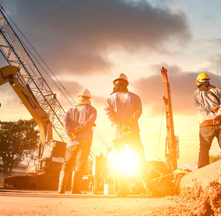 Three construction workers in hard hats stand near heavy machinery at a construction site during sunset.